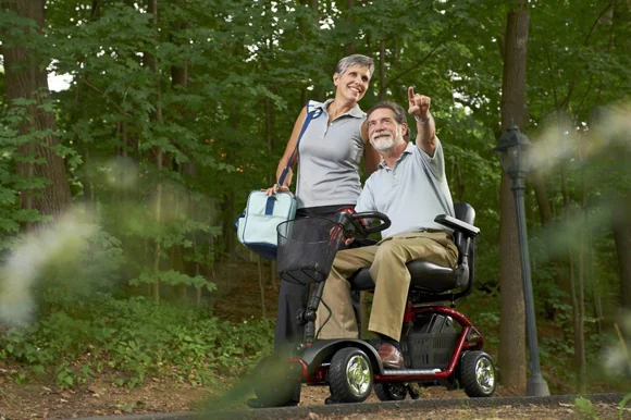 Smiling senior couple on a shaded park path, one seated on a four-wheel red mobility scooter, in Palm Beach County, FL